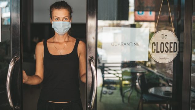 Shop owner in facemask standing at partially open shop doorway, part-time furlough concept